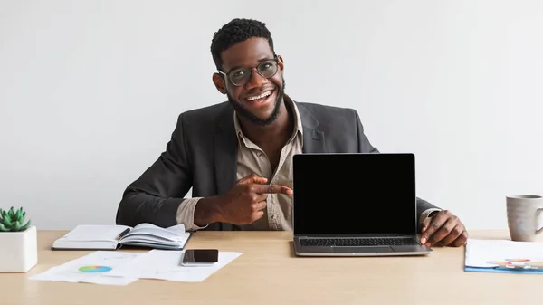 Feliz joven hombre de negocios negro sentado en el escritorio, apuntando a la computadora portátil con pantalla en blanco en la oficina en casa, maqueta —  Fotos de Stock