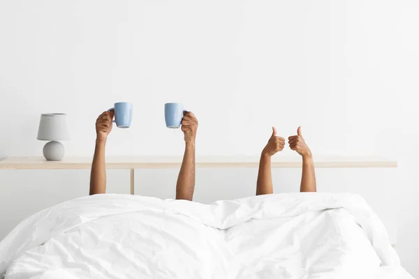 Hands of young black guy and woman stick out under blanket with thumbs up and cups of fresh coffee — ストック写真