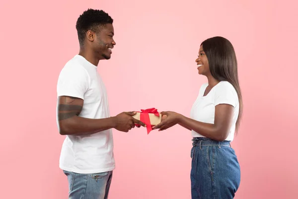 Happy black couple holding giving gift box at pink studio — стоковое фото