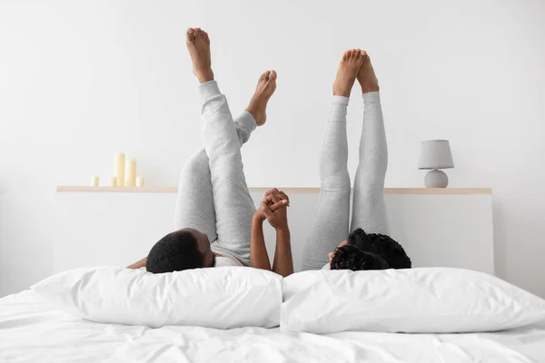 Satisfied millennial black man and woman in comfort clothes lying on bed, holding hands — Foto Stock