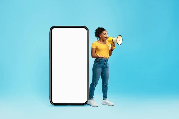 快乐的年轻黑人女人对着扩音器大喊，站在有空白屏幕的巨型智能手机旁边，模仿设计 — 图库照片