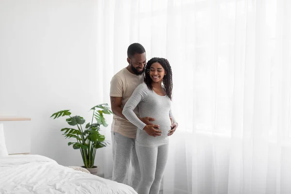 Radostně se usmívá tisíciletý africký Američan v domácím oblečení objímá svou těhotnou ženu, dotýká se břicha — Stock fotografie