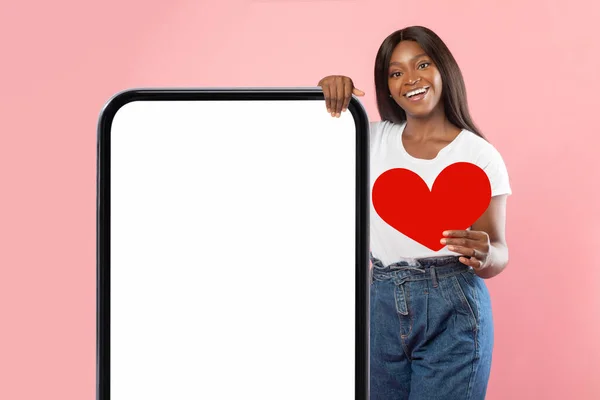 Black woman showing white empty smartphone screen holding paper heart — Photo