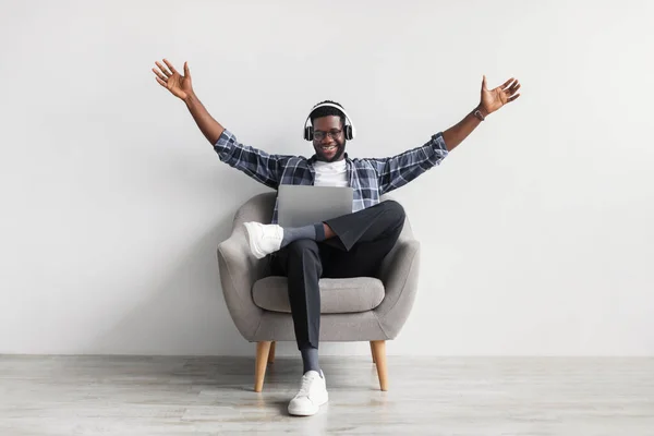 Emotional black man in headphones gesticulating, greeting someone while speaking online on laptop, sitting in armchair — 스톡 사진