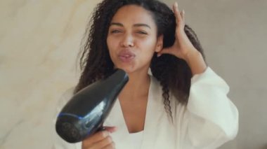 Domestic morning fun. Young positive african american woman drying her curly hair and singing into hairdryer at bathroom