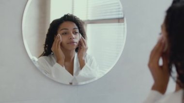 Young cute african american lady wearing bathrobe cleaning her face with moisturizing lotion and cotton pads