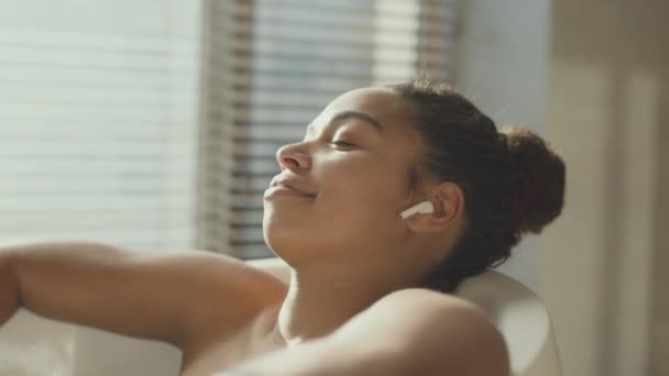 Relax time. Young african american woman relaxing in bath, listening to music with wireless earphones, tracking shot — Stockvideo
