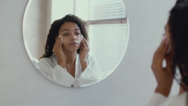 Joven linda mujer afroamericana con albornoz limpiando su cara con loción hidratante y almohadillas de algodón — Vídeo de stock