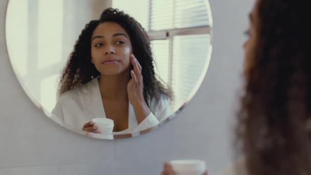 Zoom in portrait of young african american woman applying nourishing cream on her face, looking at mirror at bathroom — Video Stock