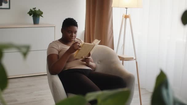 Overweight Black Female Reading Book Sitting In Chair At Home — Vídeo de Stock