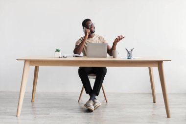 Gülümseyen genç siyahi adam cep telefonuyla konuşuyor, dizüstü bilgisayar kullanıyor, ofis masasında oturuyor, boşluğu kopyalıyor.