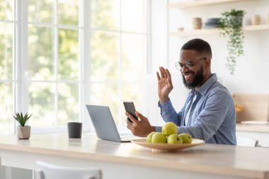 Dost canlısı, güler yüzlü, sakallı, gözlüklü siyah adam telefona el sallıyor ve dizüstü bilgisayarında çalışıyor.