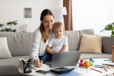 Online eğitim. Genç anne kucağında bebek, dizüstü bilgisayarla çalışıyor. Webinar izlerken not alıyor.