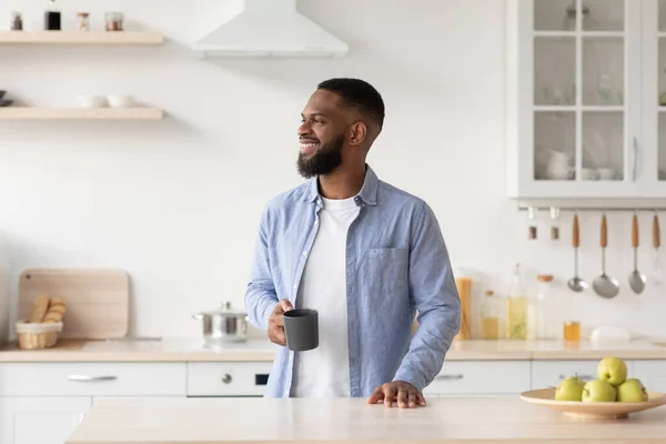 Smiling millennial black male with beard enjoys good morning and fresh coffee in cup and looks at free space — 图库照片