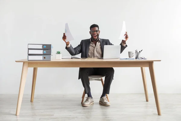 Емоційний молодий чорний чоловік з документами, що сидять за столом перед ноутбуком, відчуває себе тріумфальним, кричить в збудженні — стокове фото