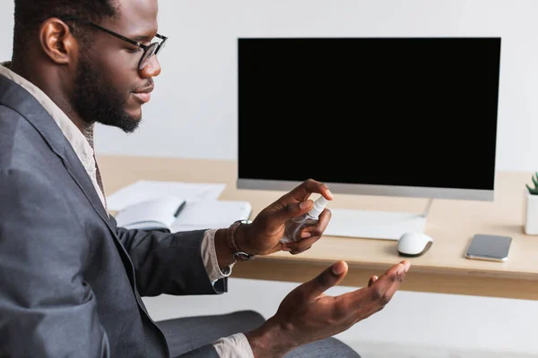 Молодий чорний бізнесмен розпилює антисептик на руках перед роботою в офісі, макет для веб-сайту на екрані монітора ПК — стокове фото