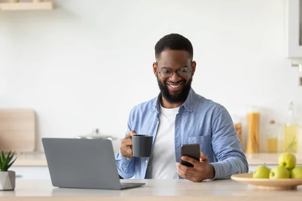 Glimlachende jonge zwarte man met baard in glazen drinkt koffie uit beker, cheques app op smartphone — Stockfoto