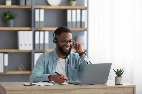Cheerful handsome millennial african american male with beard in glasses, headphones works on computer — стоковое фото