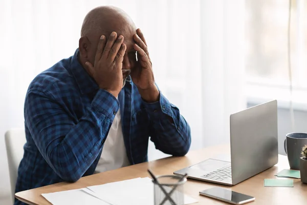 Ongelukkig senior zwart zakenman Covering gezicht zitten in de buurt van laptop Indoor — Stockfoto