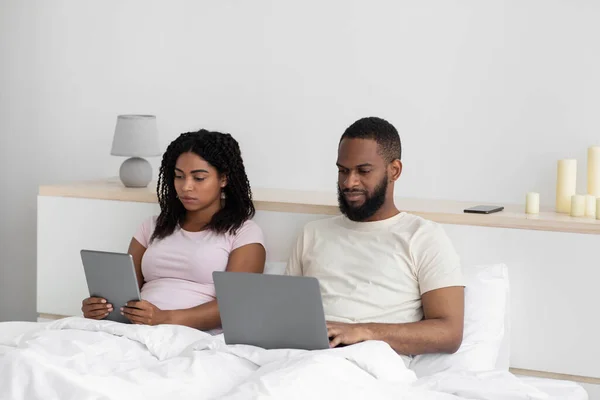 Rušné vážné tisícileté černošky manželka a manžel pracuje na notebooku a tabletu, sedí na posteli — Stock fotografie