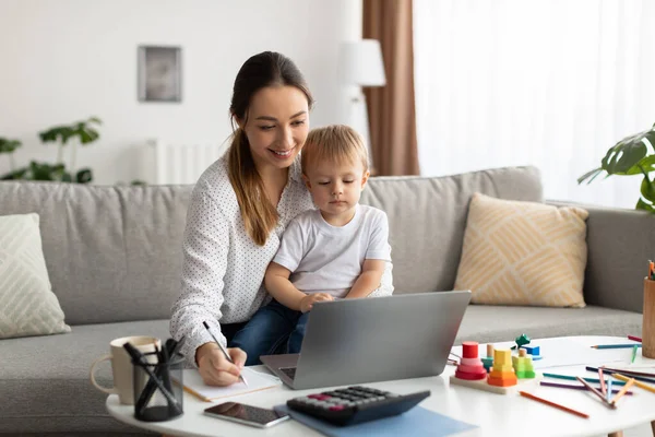 Online education. Young mother holding baby and studying with laptop, taking notes to notepad while watching webinar — Fotografia de Stock