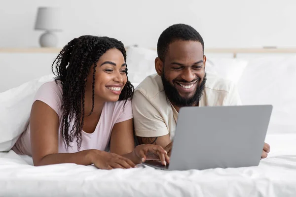 Glad smiling loving millennial black male and female lie on bed and watch video on laptop in bedroom — Fotografia de Stock