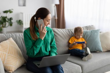 Çalışan anne. Genç bayan serbest çalışan cep telefonuyla konuşuyor ve dizüstü bilgisayar kullanıyor, oğluyla birlikte evdeki kanepede oturuyor.