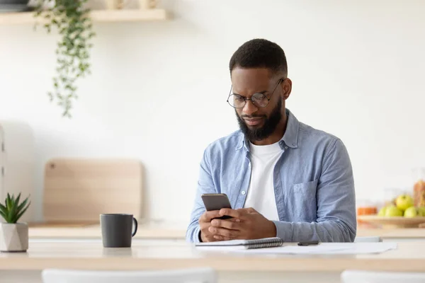 Serious young african american guy with beard in glasses chatting on smartphone works remotely — Zdjęcie stockowe