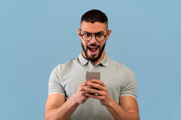 Gözlüklü genç ortadoğulu adam şok içinde telefonuna bakıyor, mavi stüdyo arka planında duruyor. — Stok fotoğraf
