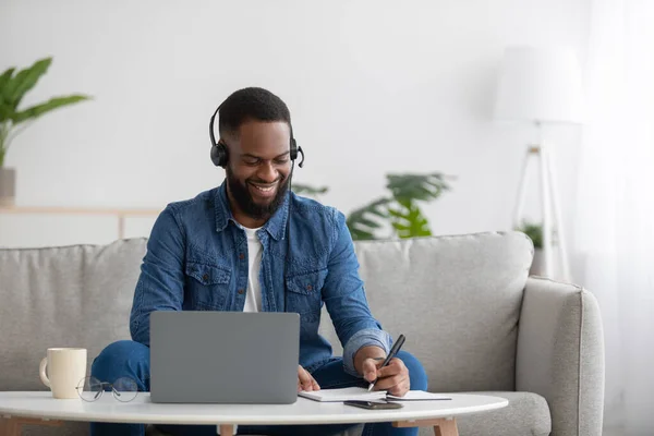 Smiling busy young black businessman freelancer or student with beard in headphones with pc making notes, study — Zdjęcie stockowe