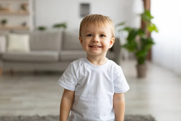 Portrait of adorable toddler boy wearing white t-shirt and smiling, posing to camera in living room interior — ストック写真