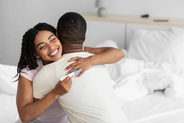 Spokojeni šťastný mladý černošky dáma objetí muž a drží těhotenský test, oslavit dobré zprávy v ložnici — Stock fotografie