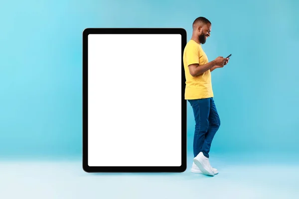 Attractive black man standing near huge digital tablet with empty screen, using smarthone, advertising new app, mockup — 스톡 사진