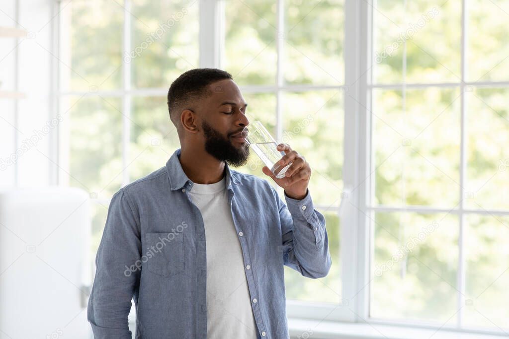 Young handsome black guy with beard drinks of clean water from glass with closed eyes at window background