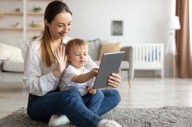 Genç anne ve küçük oğlu tabletle video görüşmesi yapıyor. Kadın web kamerasına el sallıyor ve gülümsüyor.