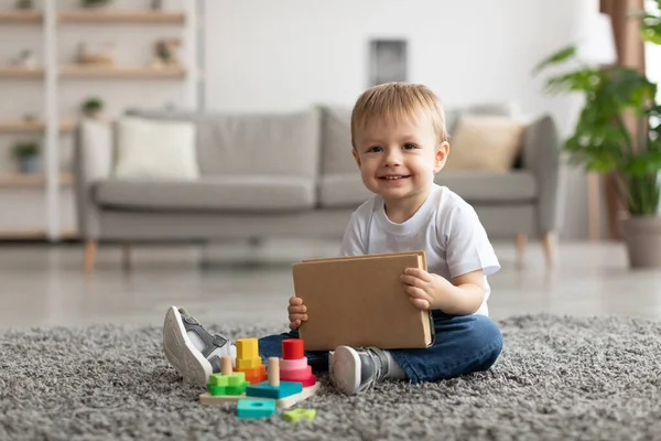 Bedårande barn pojke hålla bok medan du leker med stapling och sortering leksak, sitter på matta och ler mot kameran — Stockfoto