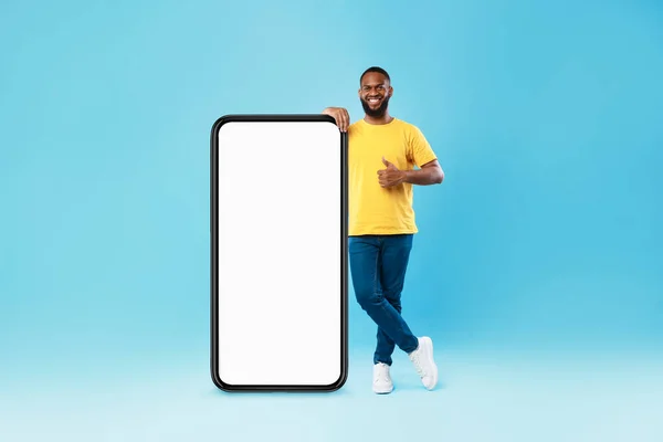 Handsome black guy standing near big cellphone with empty screen, showing thumb up, recommending cool website, mockup — Stockfoto