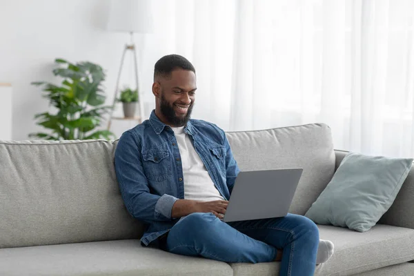 Happy young black man manager with beard sits on couch typing on pc in living room interior — Zdjęcie stockowe