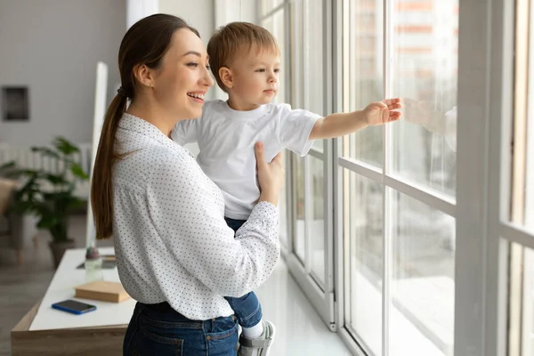 Gelukkig moeder houdt peuter zoon op handen, staande door raam toont hem de wereld buiten, jongen wijst naar straat — Stockfoto