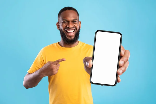 Handsome happy black guy pointing at smartphone with blank screen on blue background, mockup for website or mobile app — Fotografia de Stock