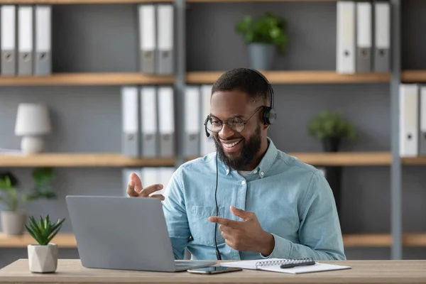 Smiling young black guy teacher or student with beard in glasses and headphones shows his fingers at pc screen — Zdjęcie stockowe