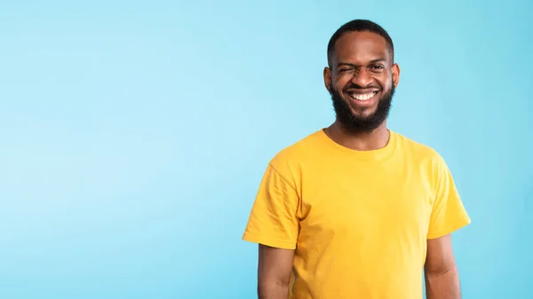 Happy bearded young black man in yellow t-shirt looking at camera and winking on blue background, banner with copy space —  Fotos de Stock