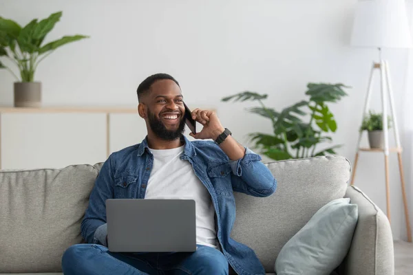Amistoso joven empresario afroamericano alegre con barba se sienta en el sofá con PC hablando por teléfono — Foto de Stock