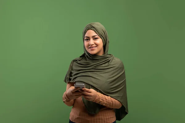 Cheerful muslim lady in hijab holding smartphone and smiling at camera, standing over dark green studio background — 图库照片