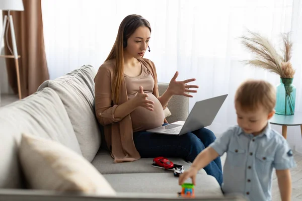 Freelancer pregnant mother working on laptop remotely from home, having video call, child boy playing near mom — Photo