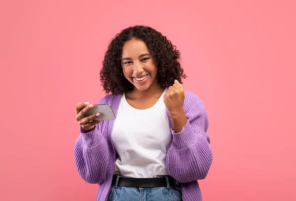 스마트폰을 들고 눈에 띄게 흥분 한 젊은 흑인 여성 이 핑크 배경으로 온라인 복권에 당첨됐다. — 스톡 사진