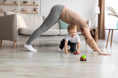 Genç anne, küçük oğluyla yoga yapıyor. Çocuk oyuncaklarla oynarken köpek pozisyonunda duruyor.