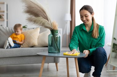 Genç anne kahve masasının tozunu alıyor ve küçük oğlu cep telefonuyla çizgi film izlerken evi toparlıyor.