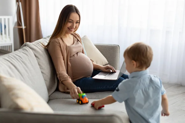Caucasian young pregnant mother working remotely on laptop with her little baby son, playing with kid and using computer —  Fotos de Stock