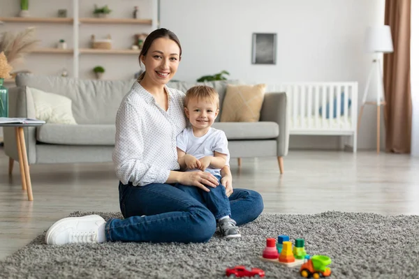 Happy caucasian mother and toddler son smiling at camera, playing together, sitting on floor carpet in living room — ストック写真